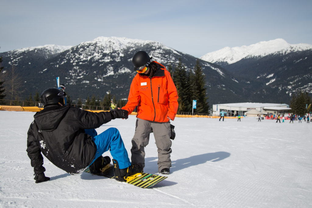 An instructor helps a snowboarder to stand up