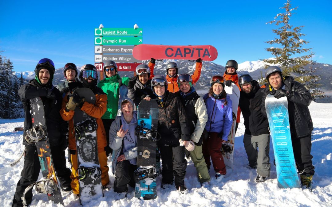 A group of young people in snowboard gear pose for a photograph with their snowboards, smiling