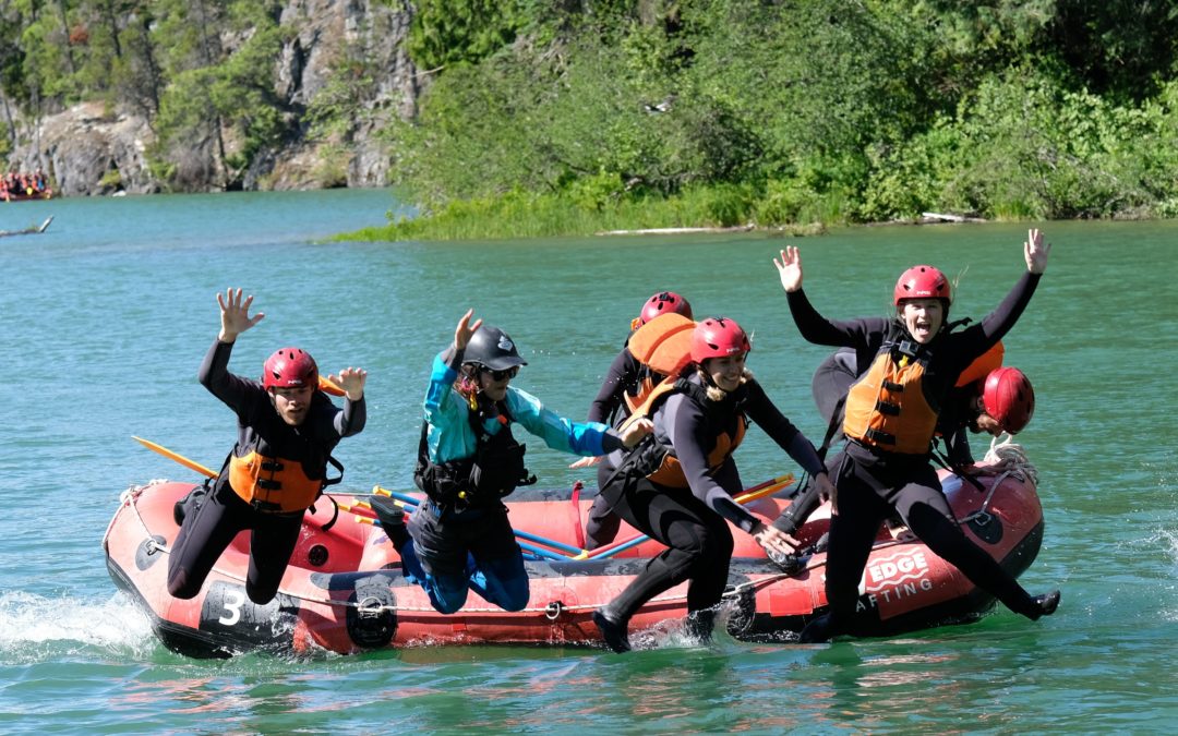 A group of people in wetsuits and life jackets jump off an inflatable raft into a clear blue lake
