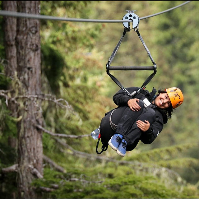 a woman rides a zipline amongst green trees. she is smiling and holds her knees to her chest in mid-air