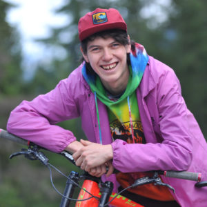 A photograph of a young man in a red hat and pink jacket. He is leaning on a mountain bike and smiling at the camera. Photo: Mike Wakefield