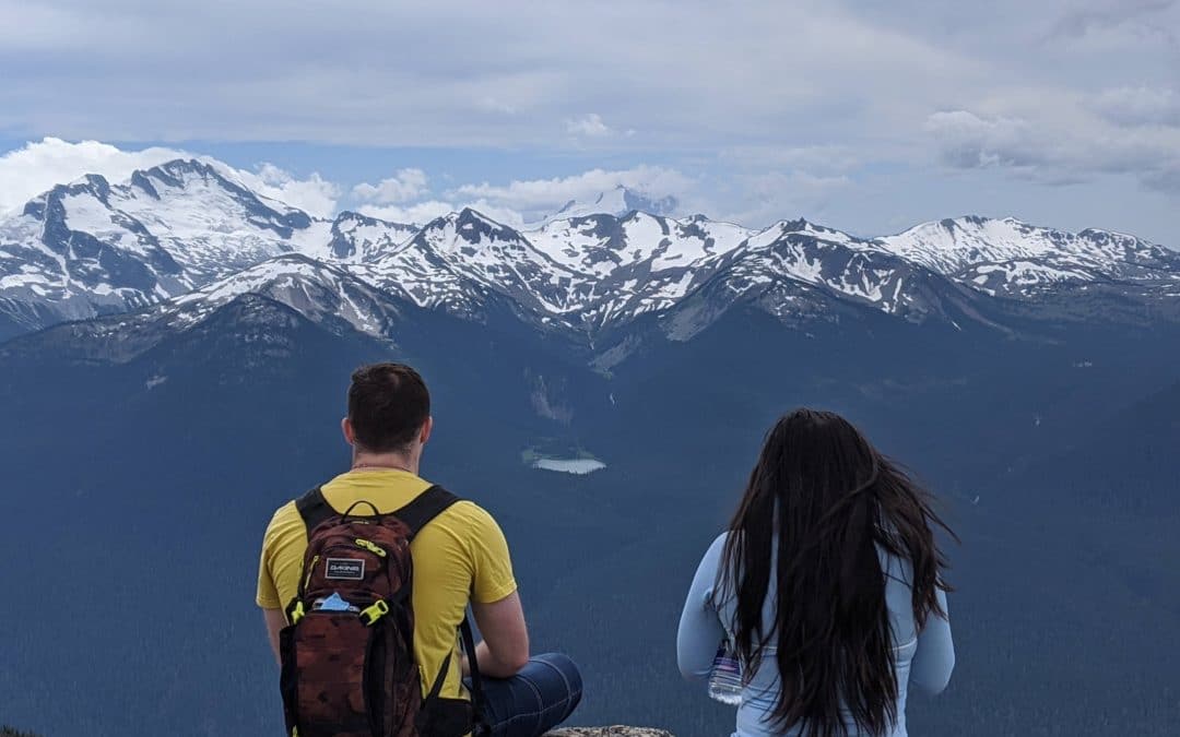 A young woman and a young man who have experienced homelessness sit looking out at a vast mountain landscape in whistler BC. Their backs are to the camera, but they appear content. They are participants in Zero Ceiling's Work 2 Live program, which was funded by the resort municipality of whistler this year to help with covid relief.