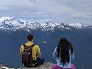 A young woman and a young man who have experienced homelessness sit looking out at a vast mountain landscape in whistler BC. Their backs are to the camera, but they appear content. They are participants in Zero Ceiling's Work 2 Live program, which was funded by the resort municipality of whistler this year to help with covid relief.