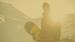 A photograph of a young man holding a snowboard and looking towards the camera. Behind him is a pristine mountain scene