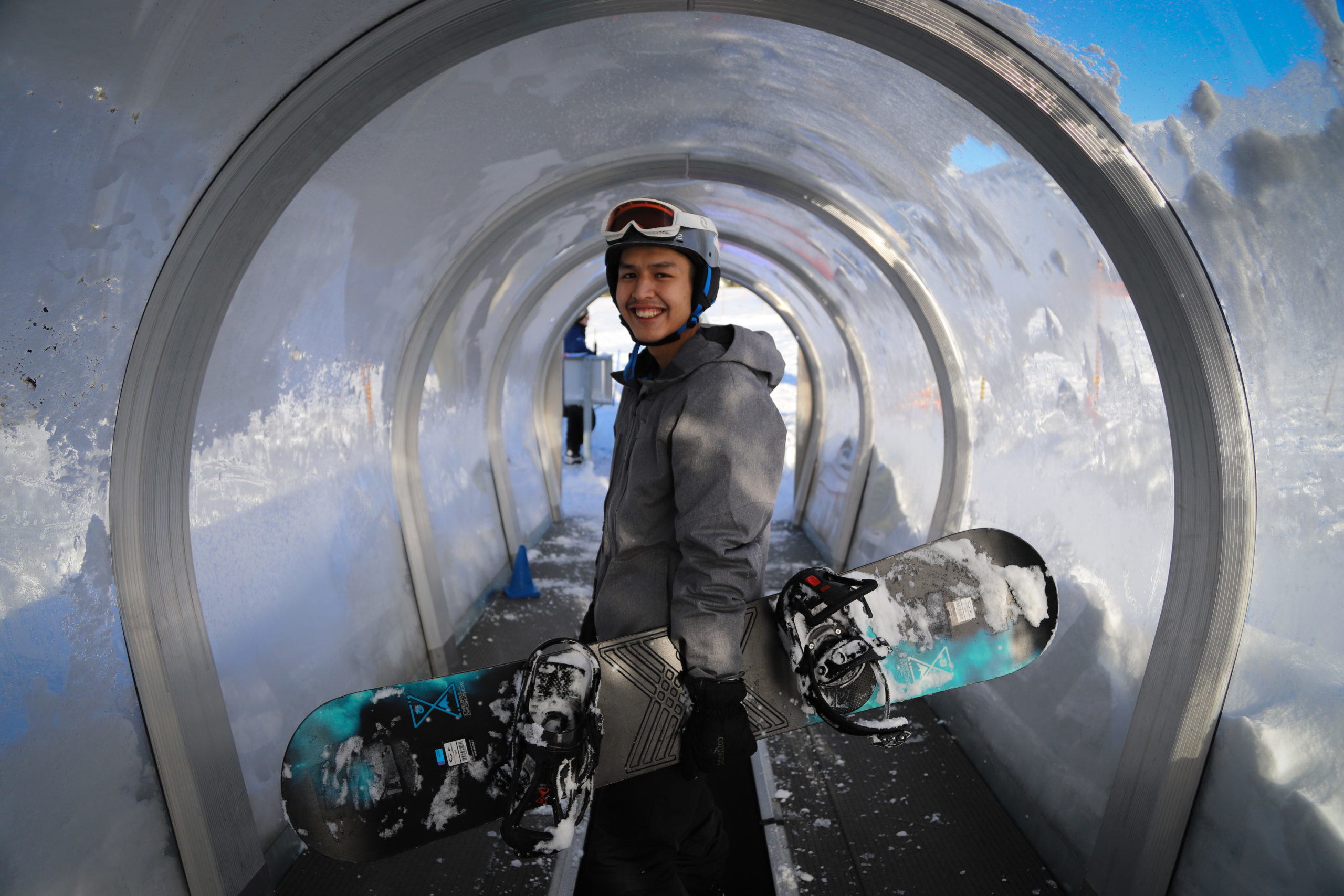 A photograph of a young Indigenous man. He is holding a snowboard and grinning widely