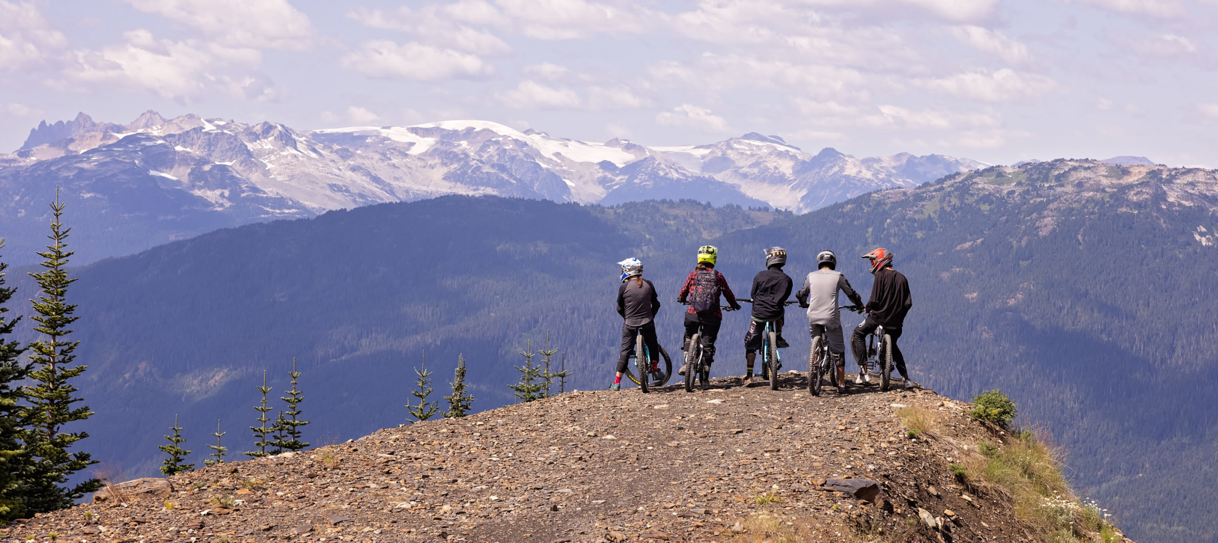 A group of young people on mountain bikes pause at a viewpoint and look out over a mountain vista. Their backs are to the camera and it is a beautiful summer day