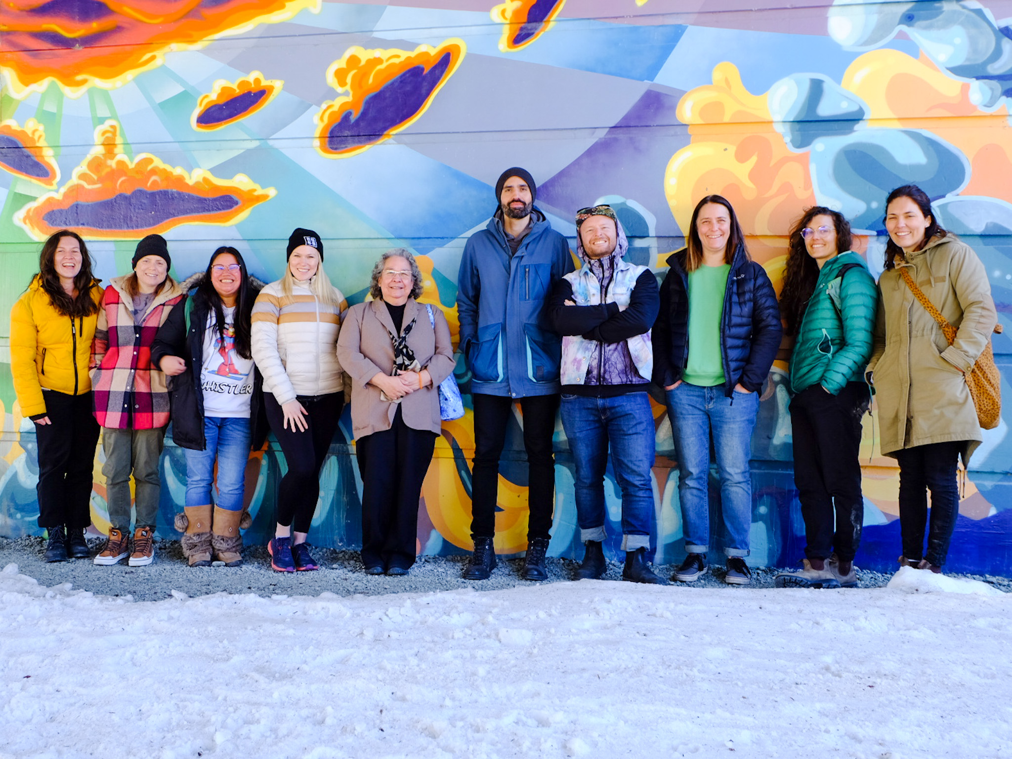 A photograph of the Zero Ceiling team. They are dressed in bright colours and standing in front of a bright mural by Chilli Thom