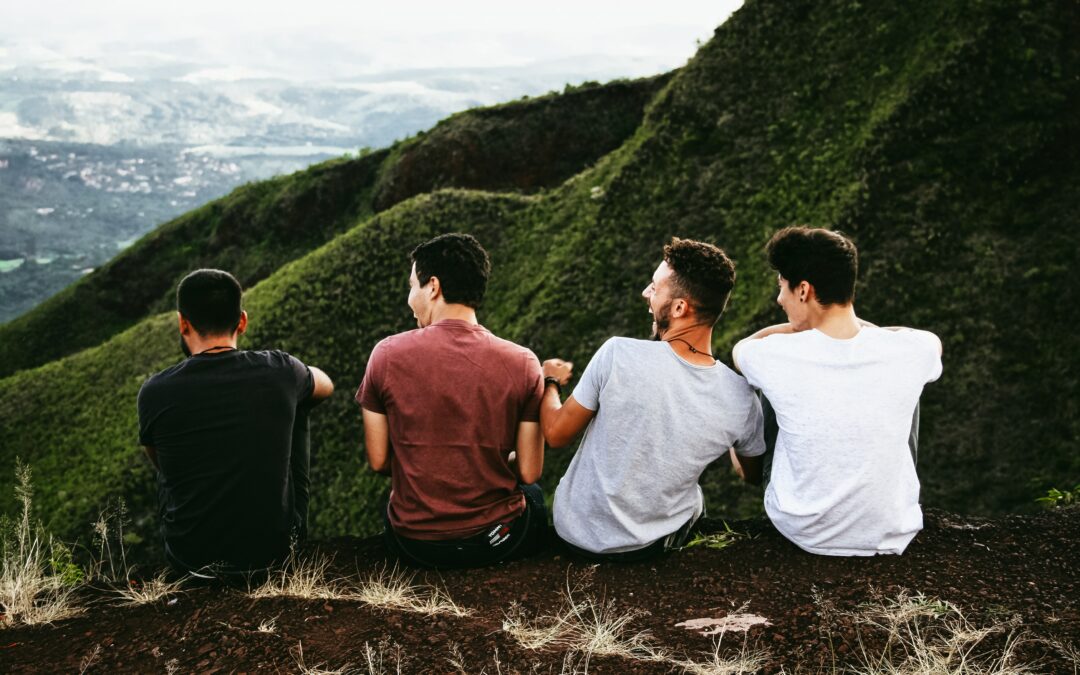 Four men laughing on the edge of a mountain with more mountain tops in the background. They're laughing with each other