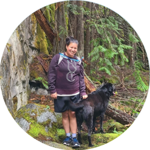 Jill is an Indigenous woman in her mid-30s. She is chubby but loves trail running, hiking, and snowboarding. She is posing with her nephew dog, Goku in a moss-covered secret riverside spot.