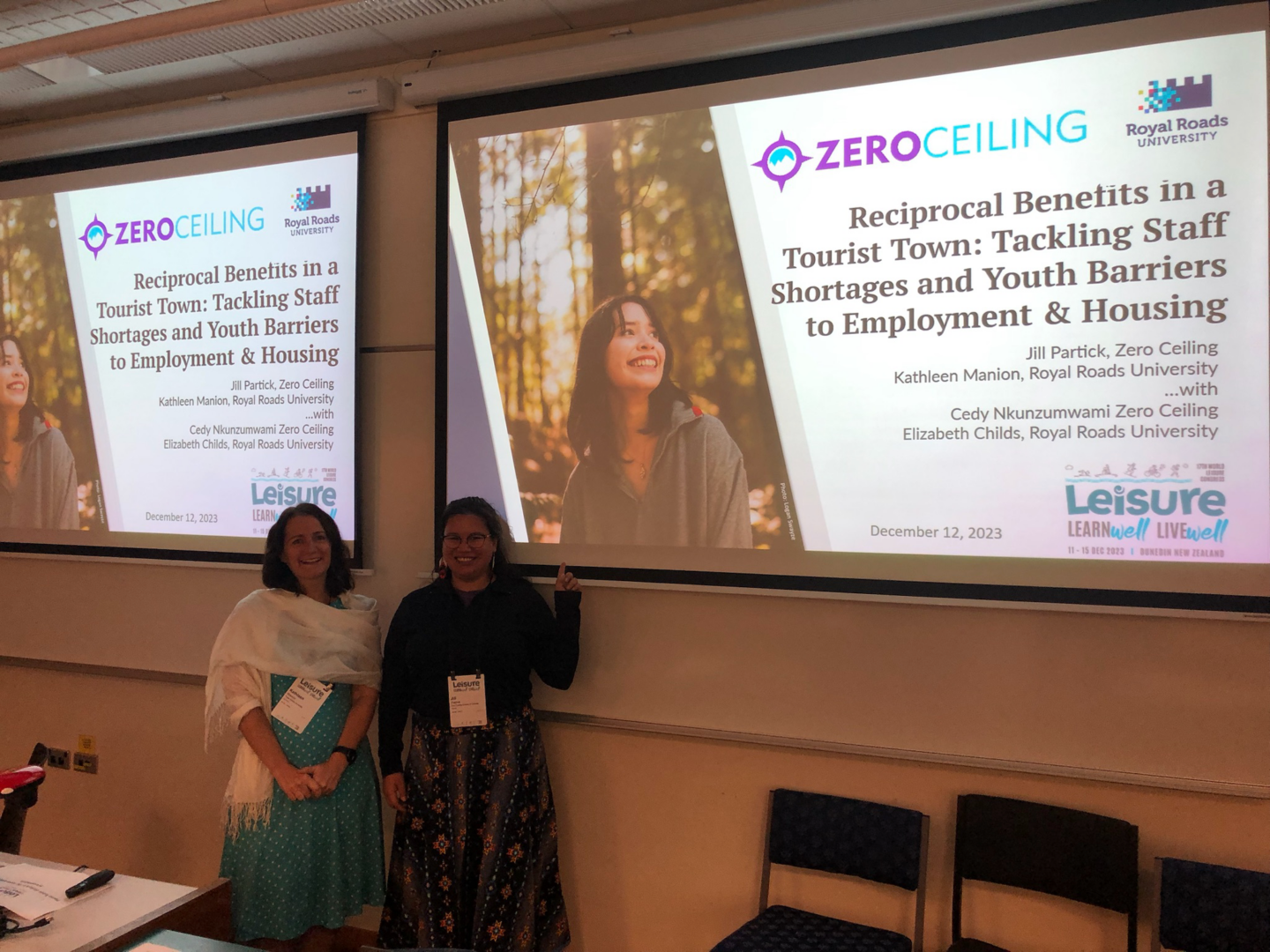 Two women stand in a classroom in front of a projection screen that shows the title page of a Powerpoint presentation.